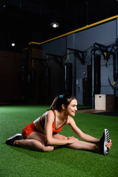 Smiling sportswoman reaching sneaker while doing stretching exercise in sport center — Stock Photo