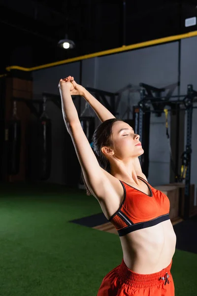 Sportive woman in sports bra stretching hands with closed eyes in gym — Stock Photo