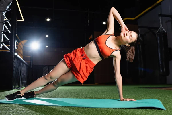 Sportiva in alto e pantaloncini che fanno esercizio laterale tavola con gli occhi chiusi in palestra — Foto stock