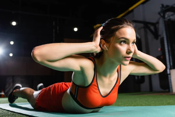 Молода спортсменка з вправами на навушниках на фітнес-маті в спортзалі, розмитий передній план — стокове фото