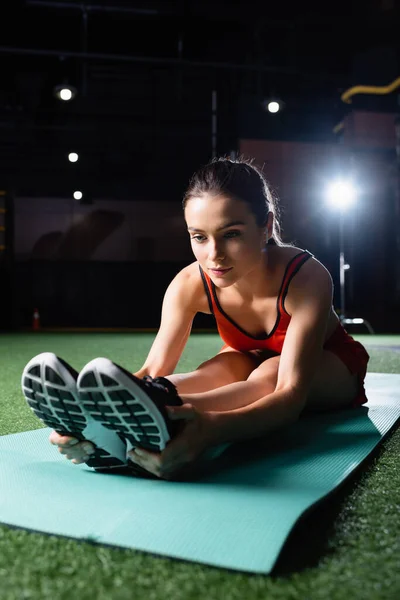 Mujer joven y deportiva tocando zapatillas de deporte mientras hace ejercicio de flexión hacia adelante sentado - foto de stock