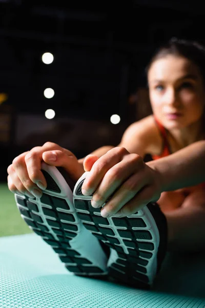 Спортсменка трогает кроссовки, делая упражнения на растяжку в тренажерном зале, размытый фон — стоковое фото