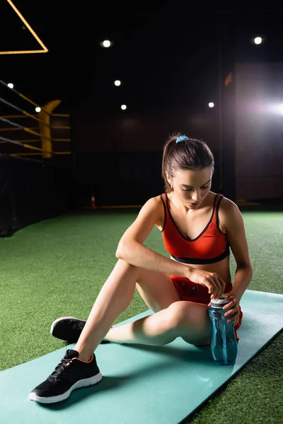 Desportista sedento abrir garrafa de esportes enquanto sentado no tapete de fitness — Fotografia de Stock