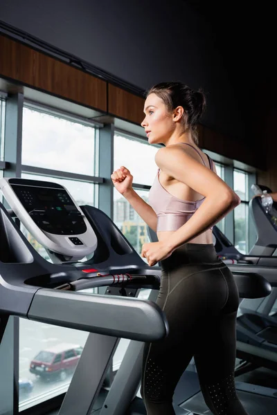 Vista laterale della sportiva in leggings distogliendo lo sguardo mentre corre sul tapis roulant — Foto stock