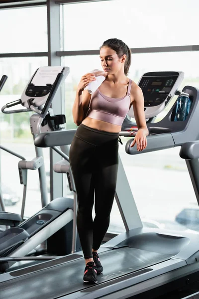 Jeune sportive en soutien-gorge et leggings debout avec serviette près de bouteille de sport sur tapis roulant — Photo de stock