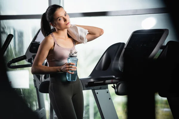 Sportive fatiguée debout sur tapis roulant, essuyant le cou avec une serviette et tenant une bouteille de sport au premier plan flou — Photo de stock