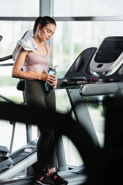 Tired sportswoman standing on treadmill with closed eyes and holding sports bottle on blurred foreground — Stock Photo