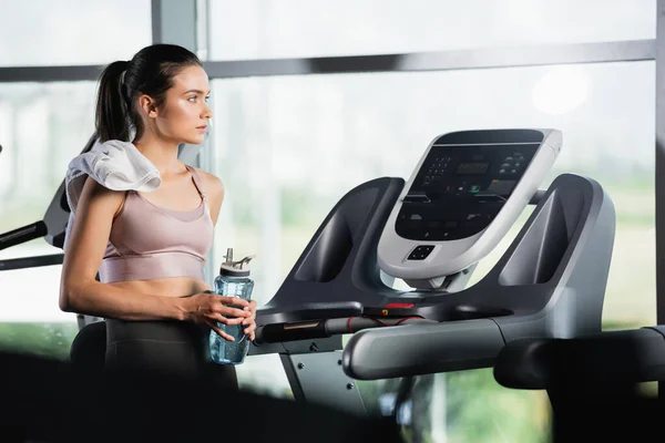 Young sportswoman looking away while standing on treadmill with sports bottle on blurred foreground — Stock Photo