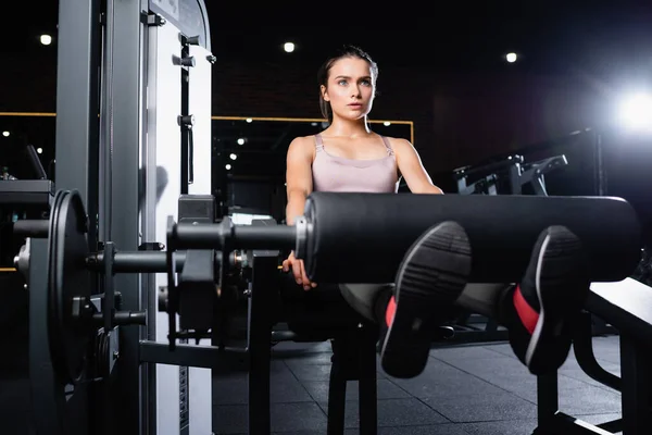 Sportswoman in bra doing leg extension exercise on training machine on blurred foreground — Stock Photo