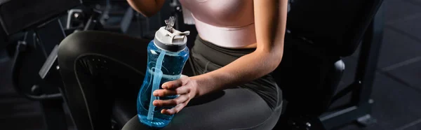 Cropped view of sportswoman sitting on training machine in gym and holding sports bottle, banner — Stock Photo