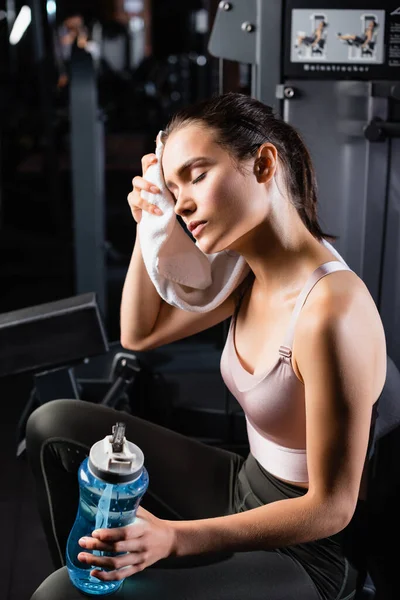 Exhausted sportswoman wiping face with towel while sitting on training machine and holding sports bottle — Stock Photo