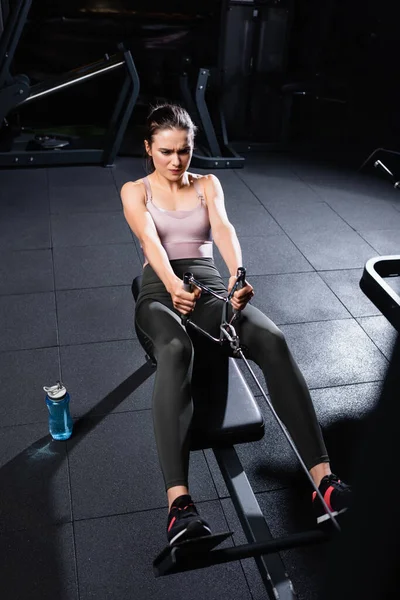 High angle view of athletic sportswoman exercising on rowing machine near sports bottle on floor — Stock Photo
