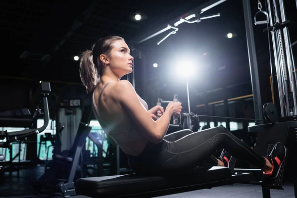 Joven deportista mirando hacia otro lado mientras hace ejercicio en la máquina de remo en el gimnasio - foto de stock