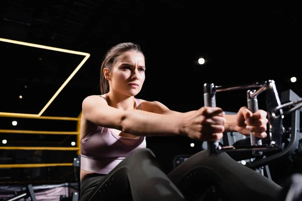 Sportive woman training on rowing machine in sports center on blurred foreground — Stock Photo