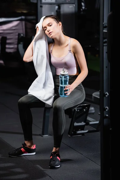 Tired sportswoman wiping head with towel while sitting on training machine with sports bottle — Stock Photo