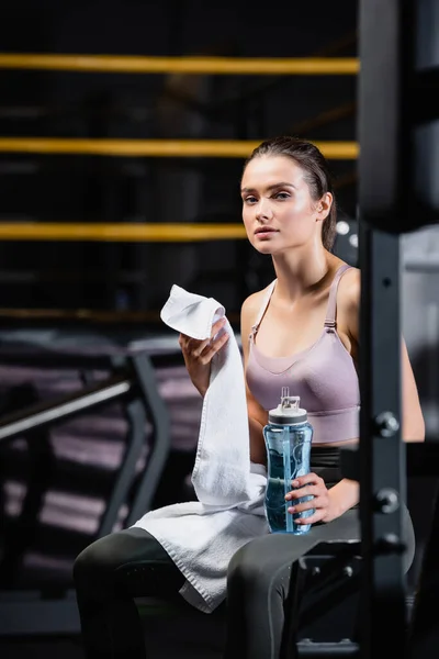 Joven deportista sosteniendo botella de deporte y toalla mientras está sentado en la máquina de entrenamiento sobre fondo borroso - foto de stock