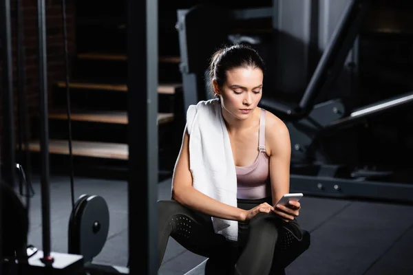 Sportlerin mit Handtuch plaudert auf Smartphone, während sie auf Trainingsgerät im Sportzentrum im unscharfen Vordergrund sitzt — Stockfoto