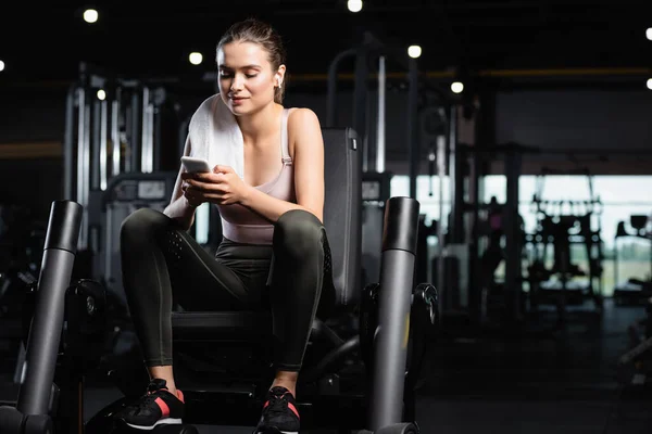 Sportswoman sitting on training machine, chatting on smartphone and listening music in wireless earphone — Stock Photo