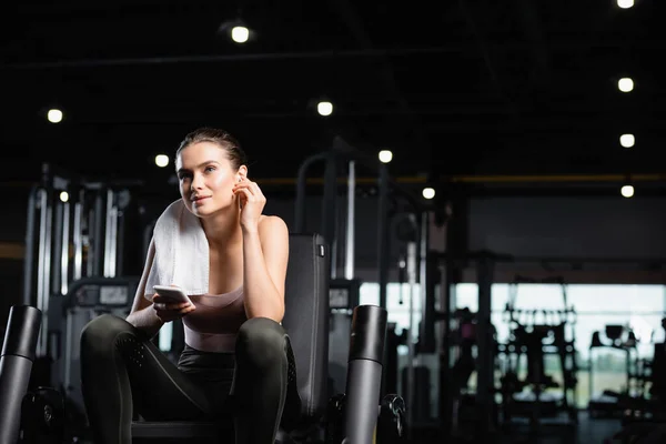 Sportive souriante écoutant de la musique dans un écouteur sans fil tout en tenant un smartphone dans une salle de gym — Photo de stock