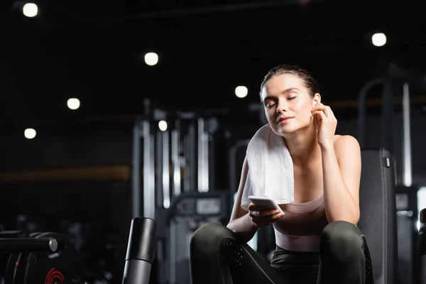 Young sportswoman listening music in wireless earphone with closed eyes while holding smartphone in gym — Stock Photo