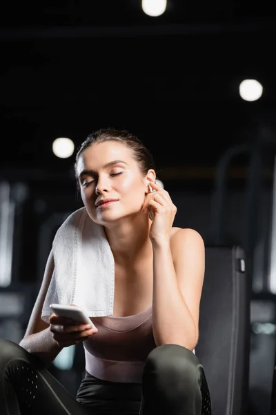Smiling sportswoman holding mobile phone and listening music in wireless earphone in sports center — Stock Photo