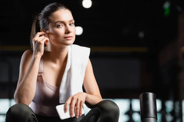 Sportswoman looking at camera while holding smartphone and listening music in wireless earphone in sports center — Stock Photo