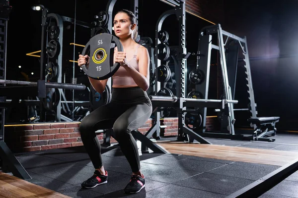 Joven deportista en leggings y zapatillas de deporte haciendo sit ups con disco de peso en el gimnasio - foto de stock