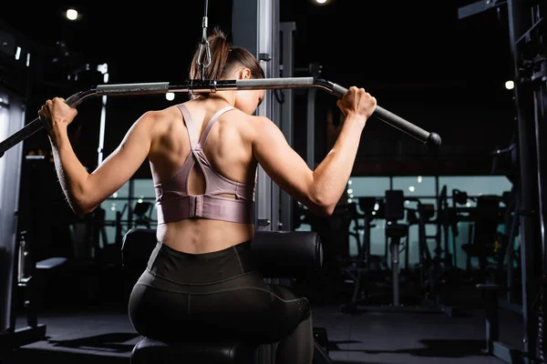 Back view of athletic sportswoman doing back and arms extension exercise on lat machine — Stock Photo