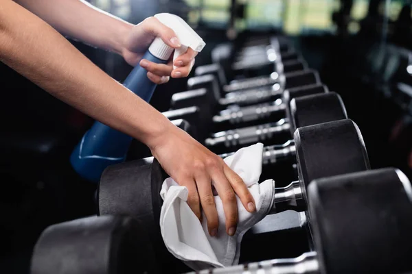 Vue recadrée de charwoman tenant le flacon de pulvérisation tout en essuyant les haltères dans la salle de gym — Photo de stock