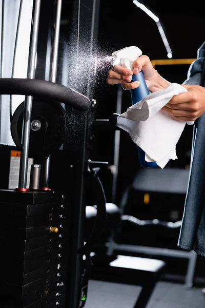 Partial view of charwoman spraying detergent on exercising machine in sports center — Stock Photo