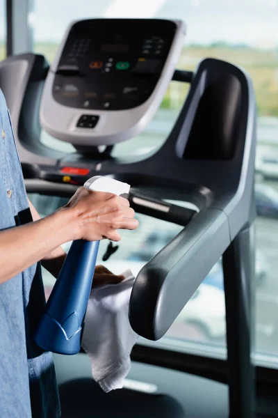 Partial view of charwoman holding spray bottle and rag while cleaning treadmill in gym — Stock Photo