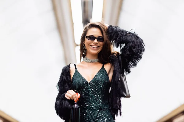 Cheerful glamour woman in black lurex dress and faux fur jacket holding wine bottle at subway station — Stock Photo