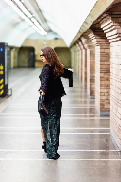 Élégante femme en robe longue noire, veste en fausse fourrure et lunettes de soleil marchant avec bouteille de vin le long de la station souterraine — Photo de stock