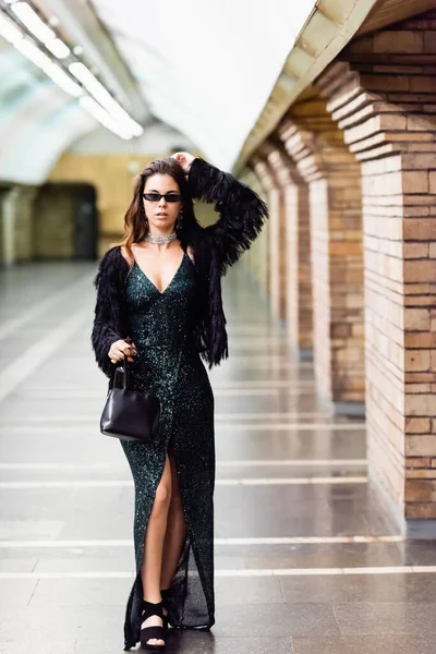 Femme à la mode en robe longue élégante et veste en fausse fourrure posant à la station de métro — Photo de stock