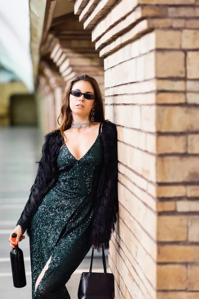 Verführerische Frau in schwarzem Lurex-Kleid und Sonnenbrille, die mit Weinflasche in der Nähe der Ziegelsäule an der Metrostation steht — Stockfoto