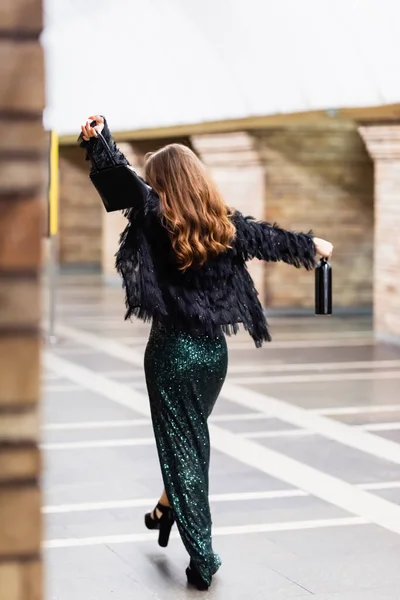 Vista posteriore di donna elegante in lungo abito lurex nero passeggiando lungo la stazione della metropolitana con borsetta e bottiglia di vino — Foto stock