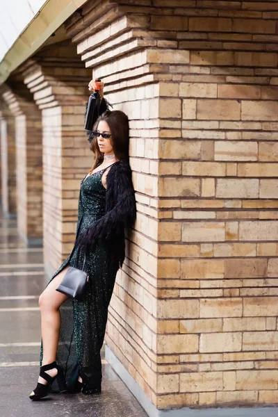 Seductive woman in long black dress leaning on brick column while holding wine bottle in raised hand — Stock Photo