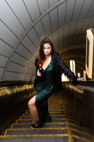 Соблазнительная женщина в черном платье с бутылкой вина смотрит в камеру на эскалаторе метро — стоковое фото