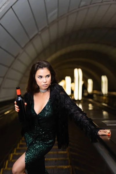 Jovem mulher elegante em vestido lurex preto segurando garrafa de vinho enquanto olha para longe na escada rolante — Fotografia de Stock