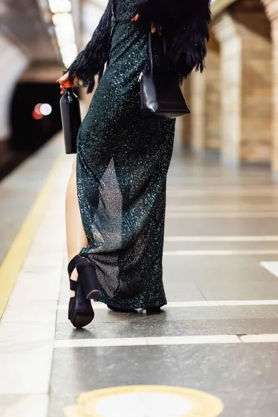 Vista cortada de mulher elegante em vestido preto longo segurando garrafa de vinho na plataforma de metro — Fotografia de Stock