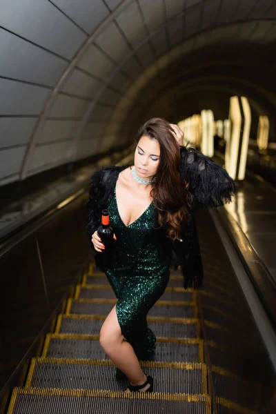Verführerische Frau im eleganten schwarzen Kleid, die das Haar berührt, während sie eine Weinflasche auf der Rolltreppe hält — Stockfoto