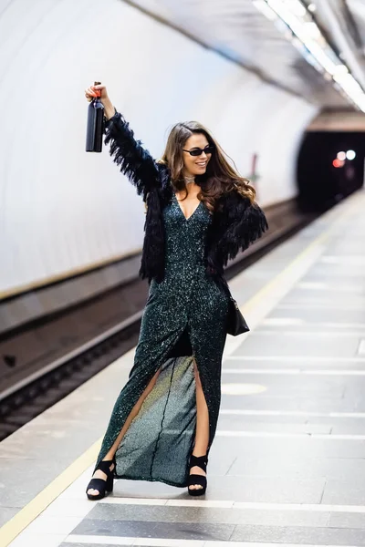Cheerful, stylish woman in black lurex dress and sunglasses holding bottle of wine in raised hand on subway platform - foto de stock