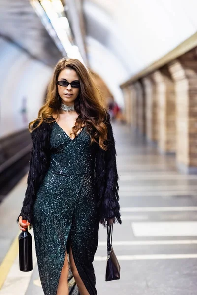 Femme séduisante en robe de lurex noir et lunettes de soleil tenant bouteille de vin sur la plate-forme de métro — Photo de stock