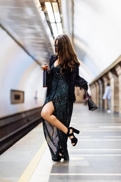 Гламурна жінка в чорній сукні люрекса, що тримає пляшку вина, позуючи на платформі метро — стокове фото