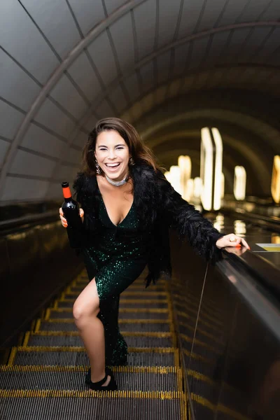 Excited woman in elegant lurex dress laughing while holding wine bottle on escalator — Stock Photo