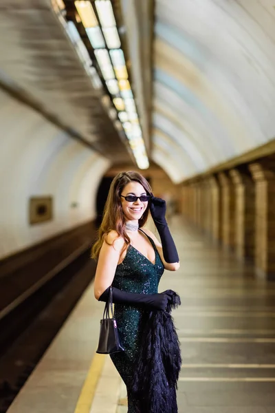 Élégante femme en robe lurex noir souriant à la caméra sur le quai de la station de métro — Photo de stock