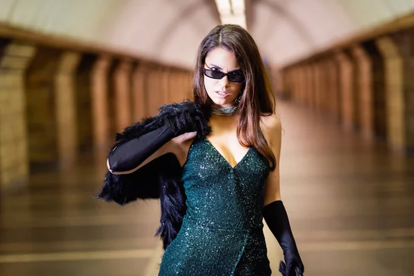 Mulher sedutora em vestido lurex preto e óculos de sol segurando casaco de pele falso enquanto posando na estação de metro — Fotografia de Stock