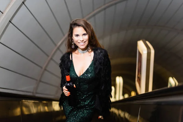 Модная женщина в черном платье улыбается в камеру, держа бутылку вина на эскалаторе — стоковое фото