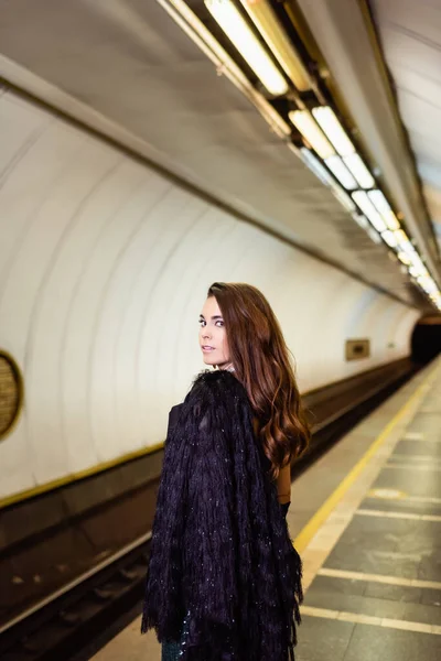 Гламурная женщина в искусственном меховом пиджаке смотрит в камеру, стоя на платформе метро — стоковое фото