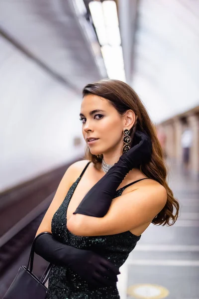 Seductive woman in elegant black dress and gloves looking at camera on subway platform — Stock Photo